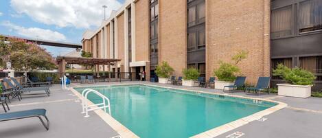 A swimming pool with lounge chairs and a covered seating area, surrounded by a brick building.