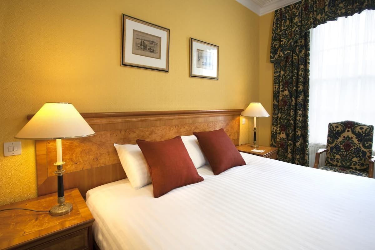 A hotel room with a bed, two bedside lamps, a wooden headboard, two framed pictures on the wall, and a patterned chair.