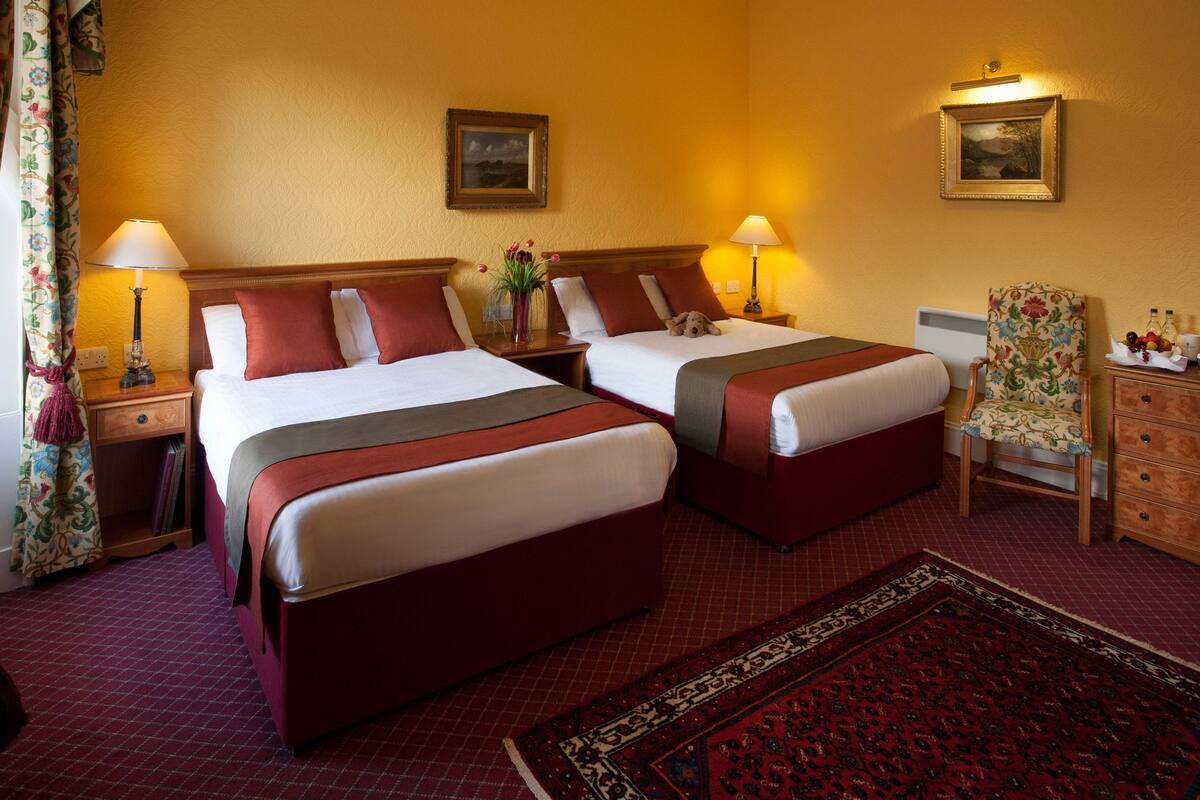 A hotel room with two beds, a wooden headboard, a floral armchair, a wooden dresser, and a red patterned rug.