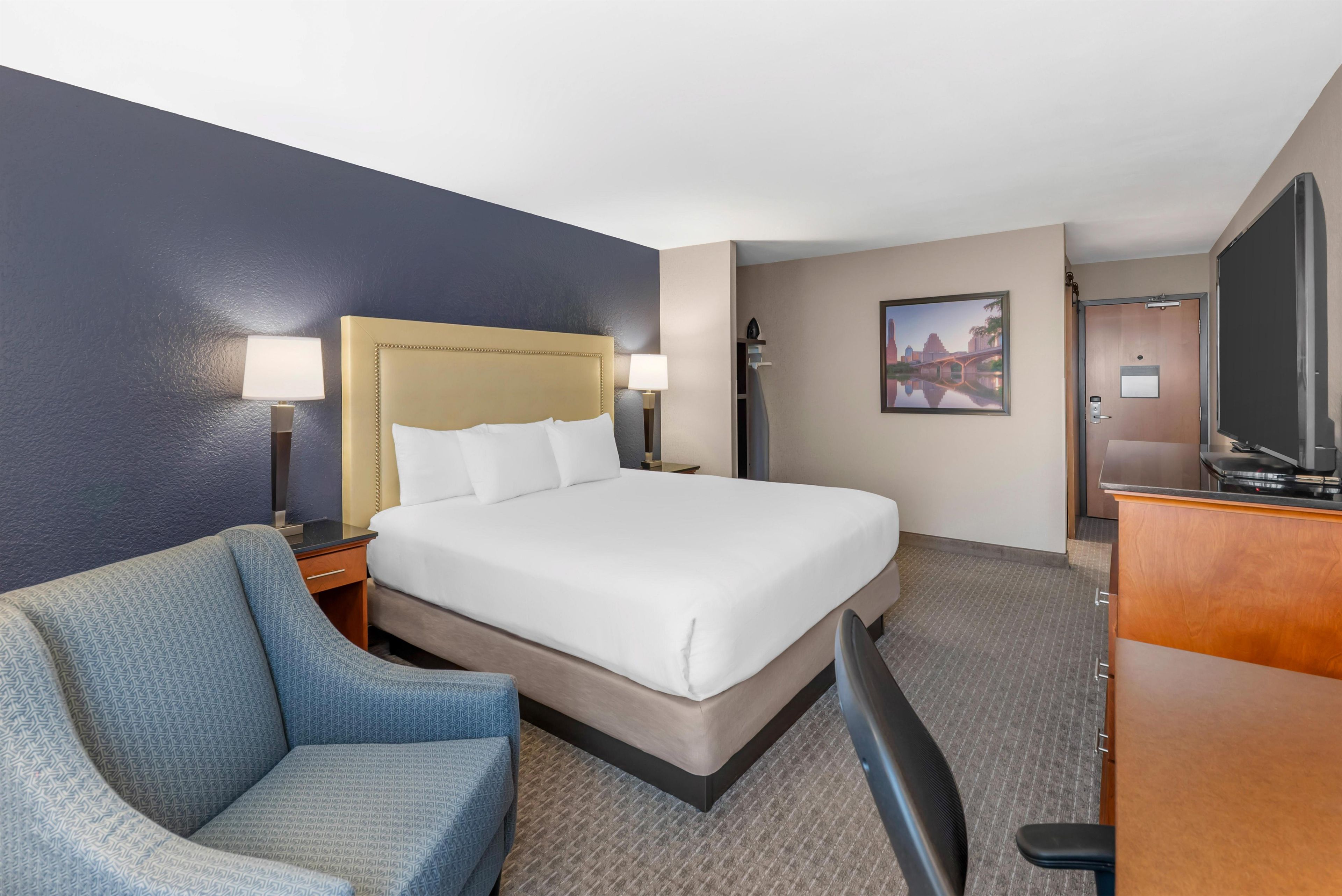 A hotel room with a bed, a chair, a desk, a television, and a painting on the wall.