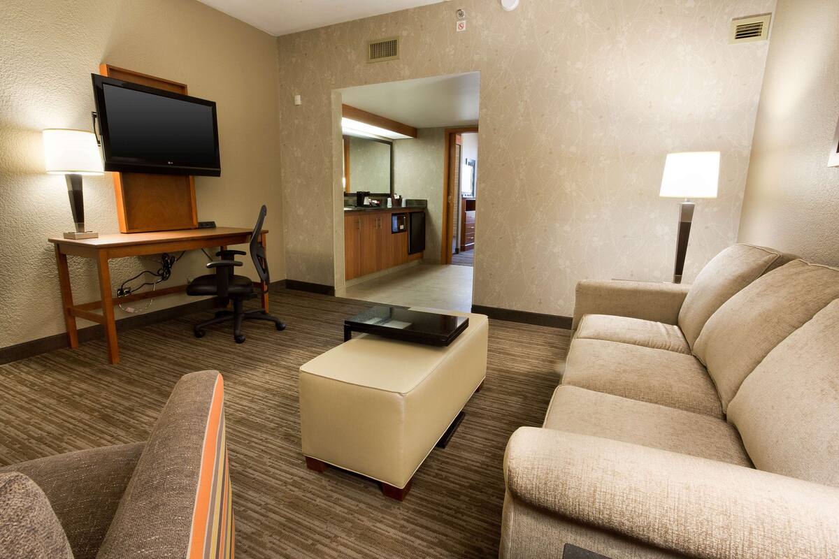 Ein Hotelzimmer mit Flachbildfernseher an einem Holztisch, einem beigen Sofa, einem kleinen Hocker und einem Sessel.