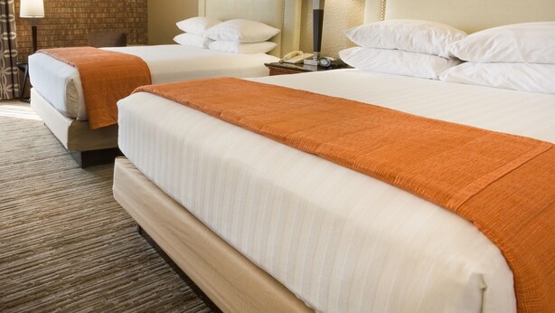 A hotel room with two beds, each with white linens and orange throw blankets. There are bedside lamps and a clock on the nightstand.