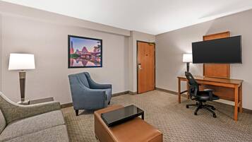 A hotel room with a sofa, armchair, desk, television, and a painting on the wall.
