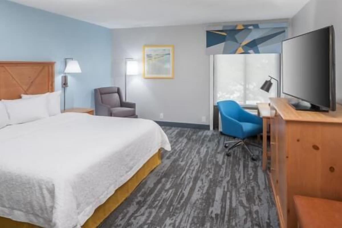 A hotel room with a bed, a desk with a computer, a chair, a TV, and a wall art piece.