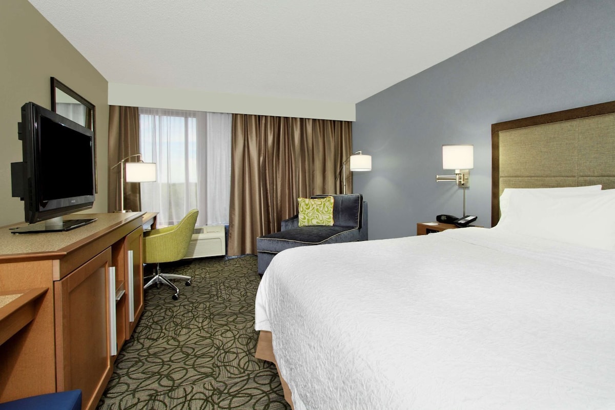 A hotel room with a bed, a desk with a TV, a chair, a lamp, and a nightstand.
