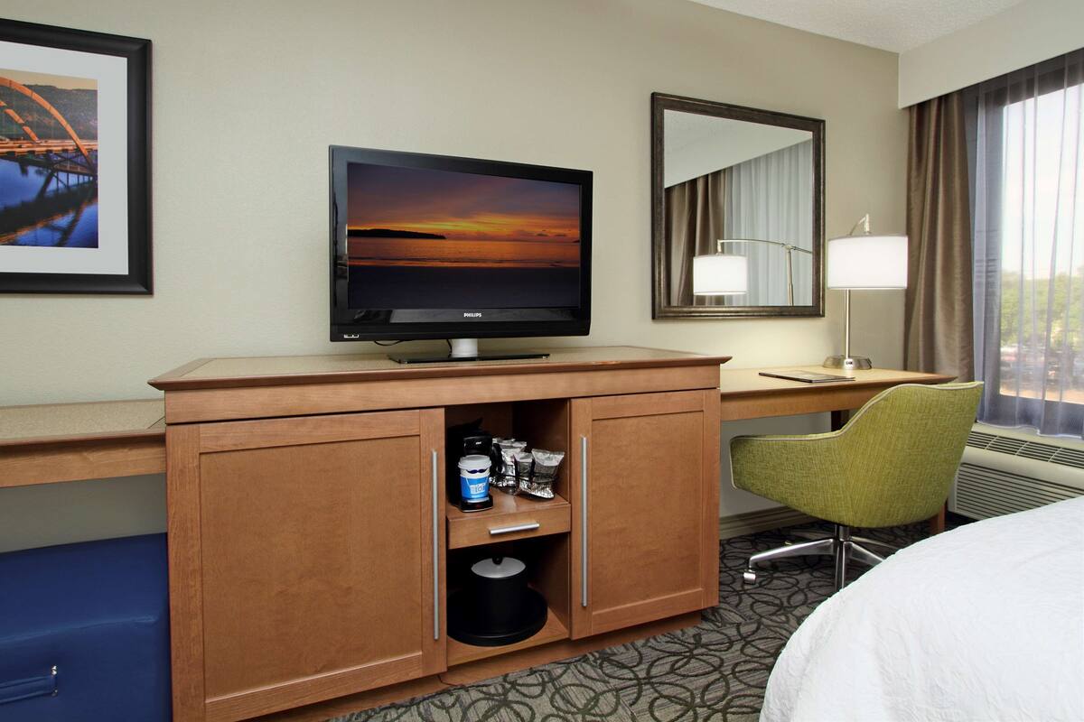 A hotel room with a wooden desk, a television, a green chair, a bed, and a large window with curtains.