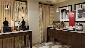 Hotelkamer met kitchenette, inclusief koffiezetapparaat, wafelijzer en magnetron. Gordijnen, een spiegel en een rode banner aan de muur.