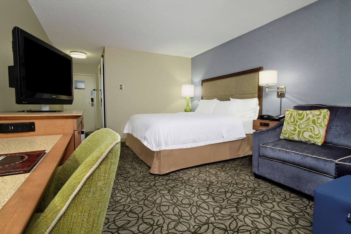 A hotel room with a bed, a desk with a flat-screen TV, a sofa, and a patterned carpet.