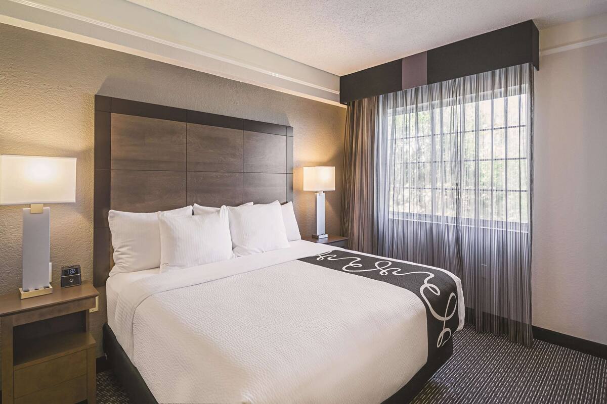 Una habitación de hotel con una cama grande, dos lámparas de noche, una mesita de noche y una ventana con cortinas.
