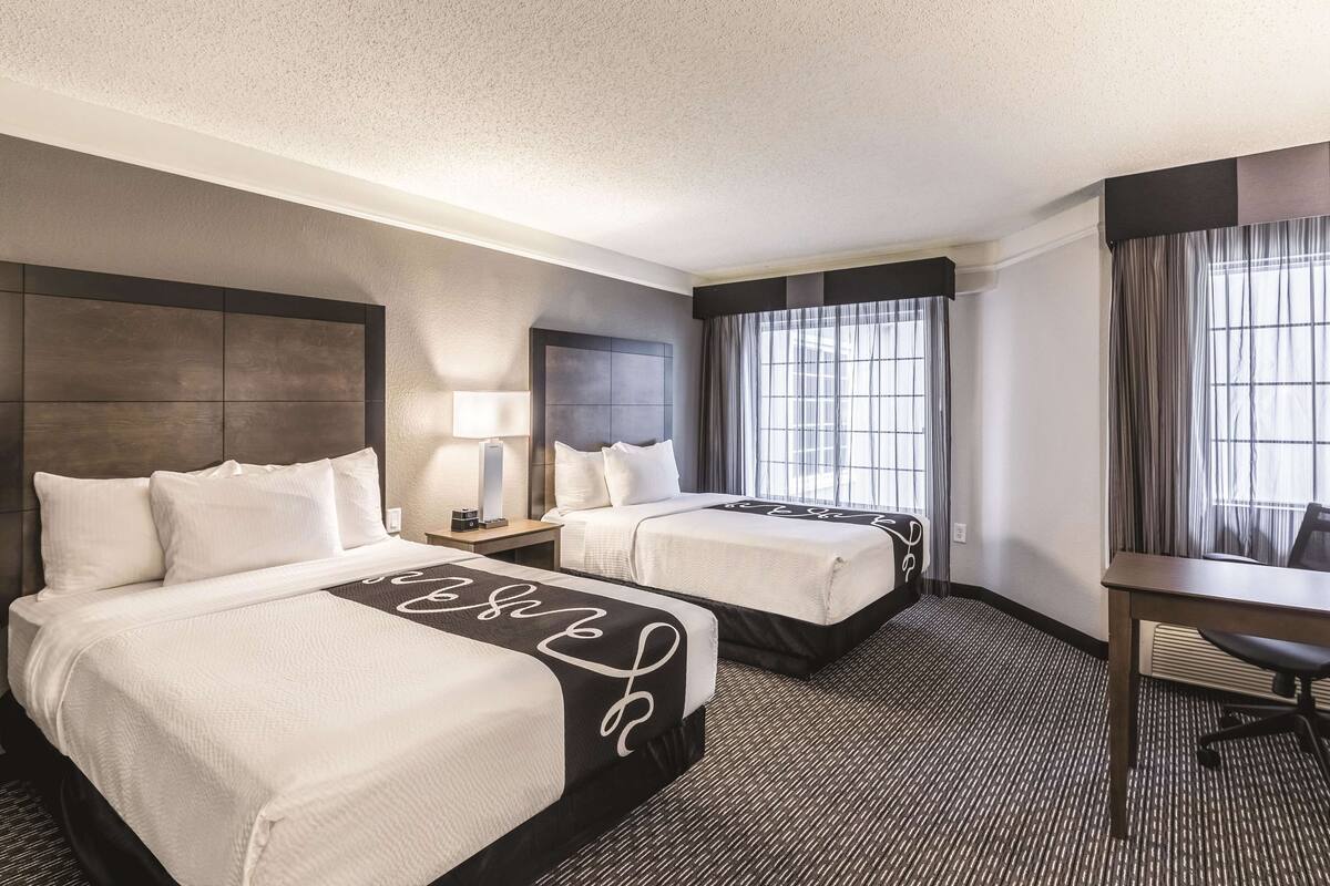 Habitación de hotel con dos camas, un escritorio, una silla, una lámpara y un amplio ventanal con cortinas.