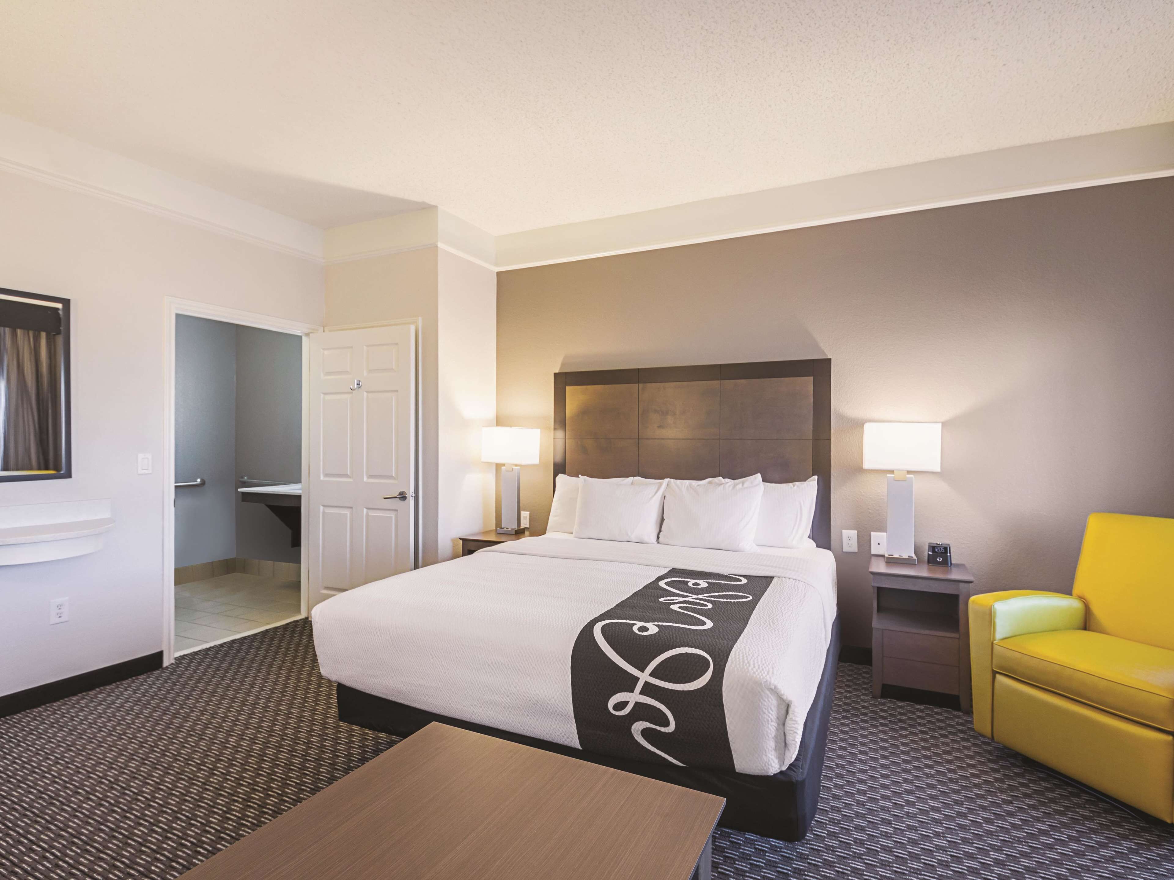 A hotel room with a large bed, two bedside lamps, a small table, a yellow armchair, and a door leading to a bathroom.