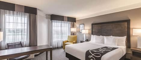 Quarto de hotel com uma cama grande, uma escrivaninha, uma cadeira e duas janelas com cortinas.