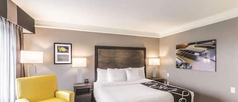 A hotel room with a bed, a yellow armchair, two bedside tables with lamps, and framed pictures on the walls.