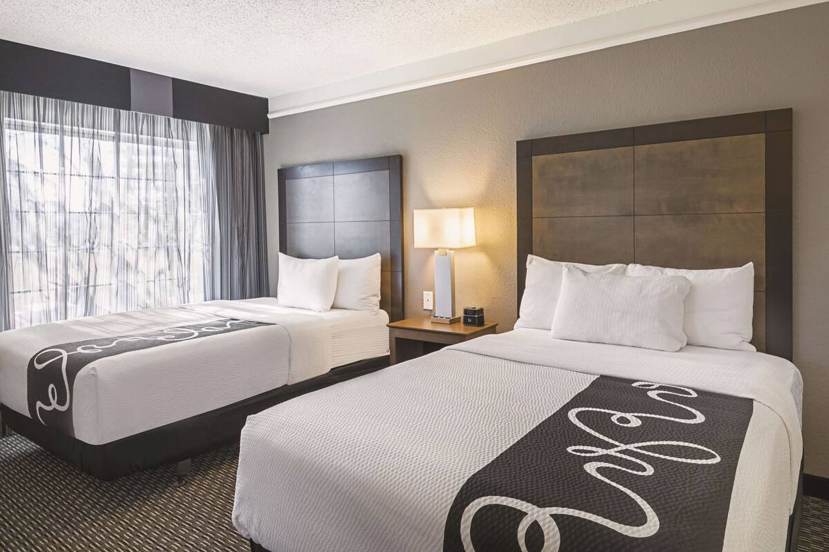 Una habitación de hotel con dos camas, un ventanal con cortinas, una mesita de noche con lámpara y un cabecero con diseño texturizado.