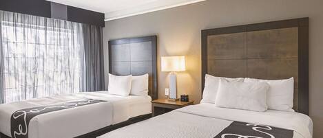 A hotel room with two beds, a large window with curtains, a nightstand with a lamp, and a headboard with a textured design.