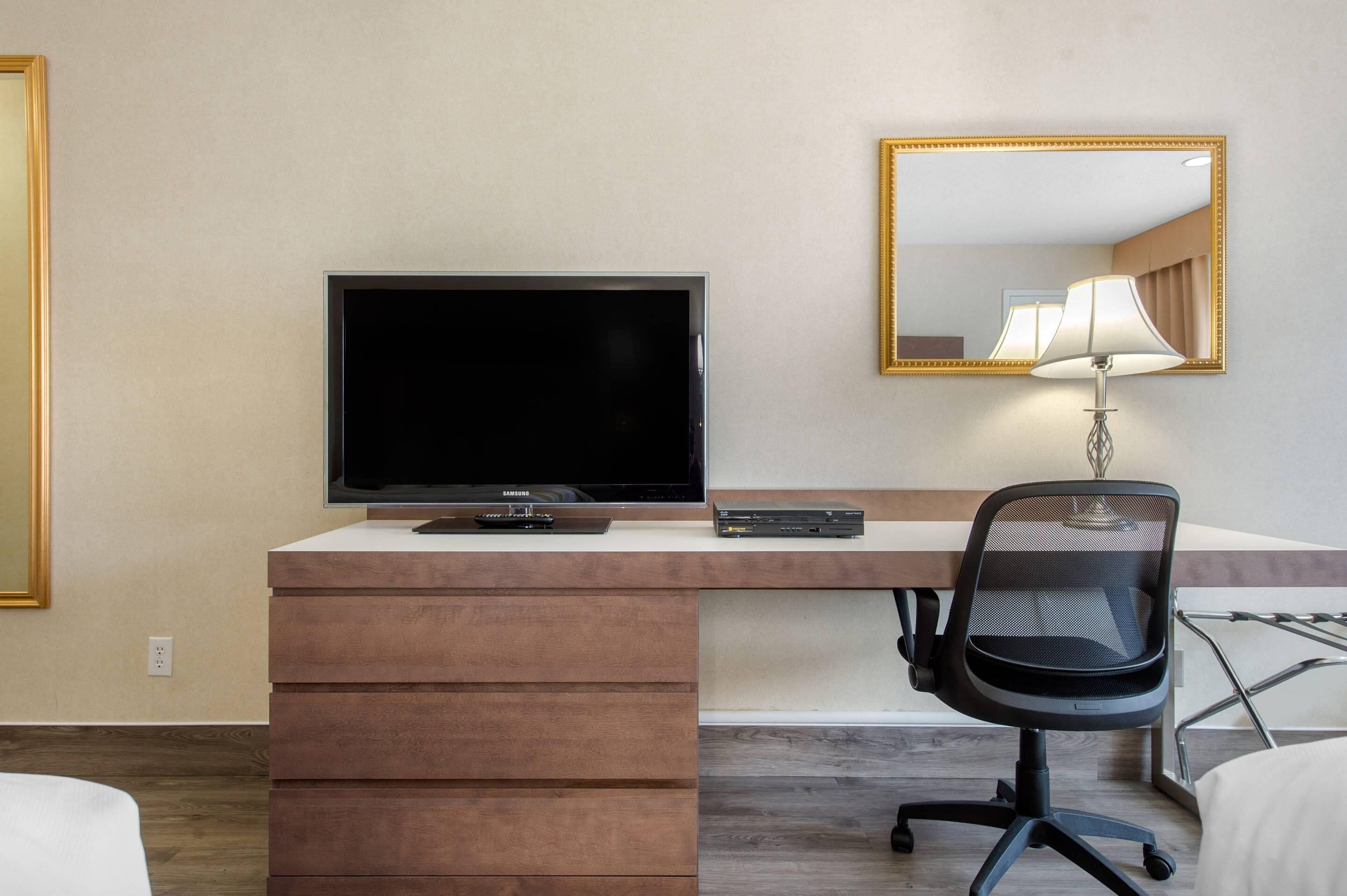 A hotel room with a flat-screen TV, a desk with drawers, a chair, and a lamp.