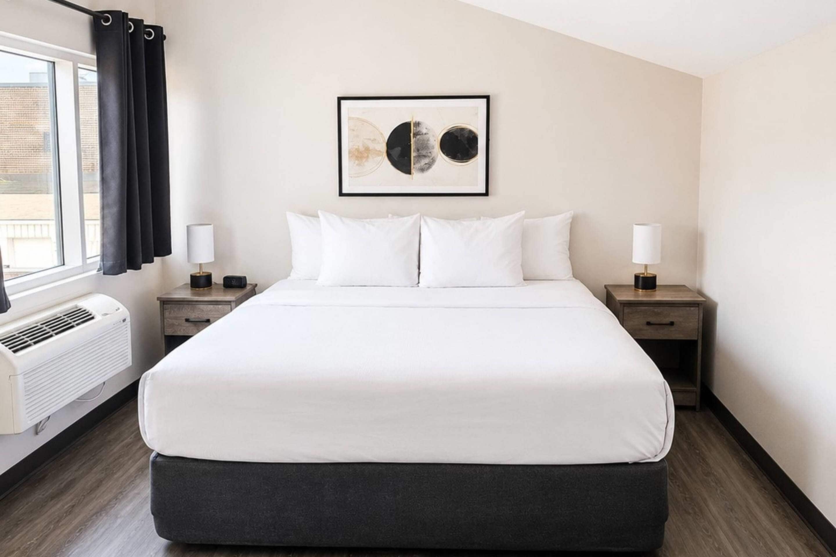 A hotel room with a large bed, two bedside tables, a window with curtains, a wall-mounted artwork, and a radiator.