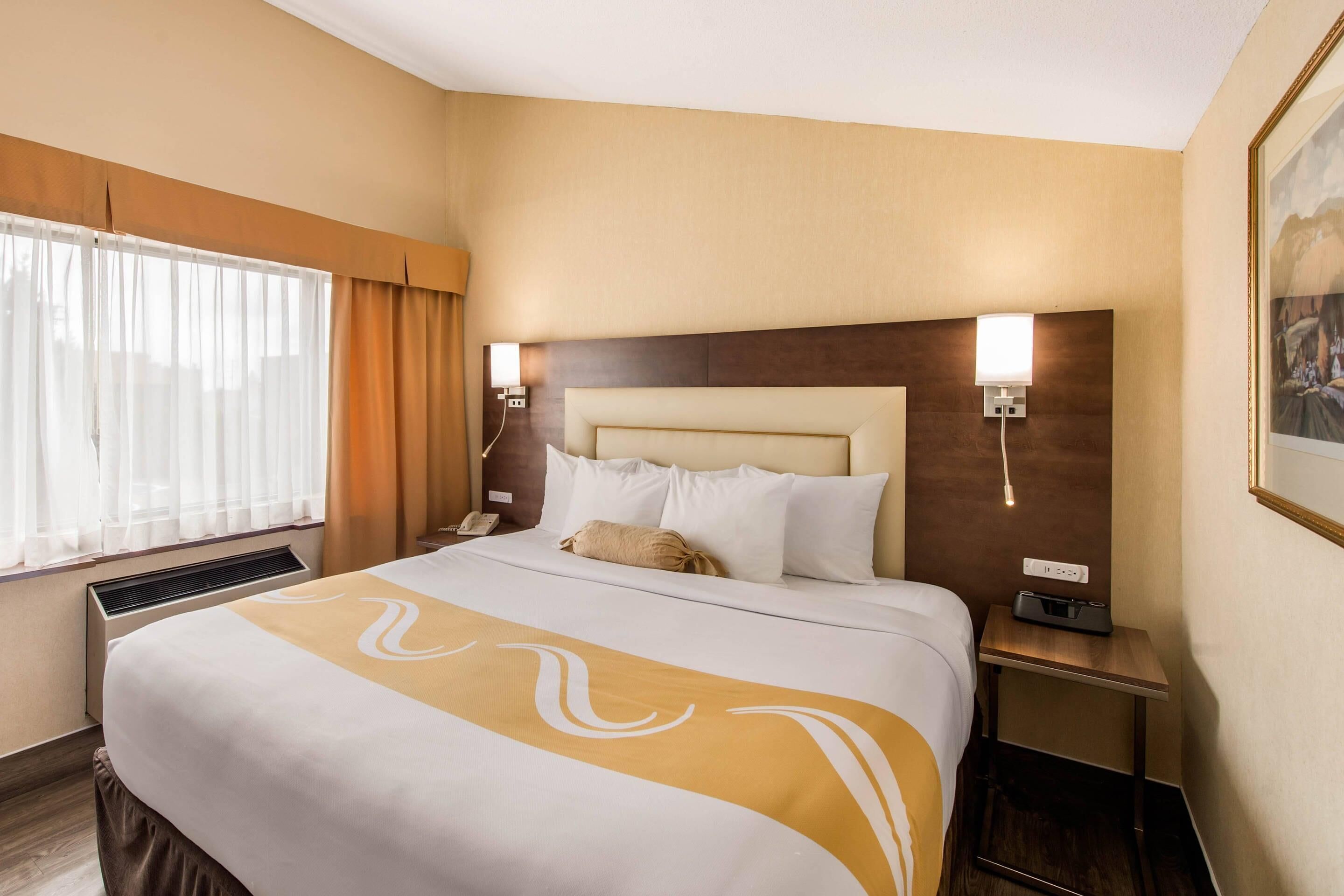 A hotel room with a large bed, a bedside table, a nightstand, a window with curtains, and a wall-mounted lamp.