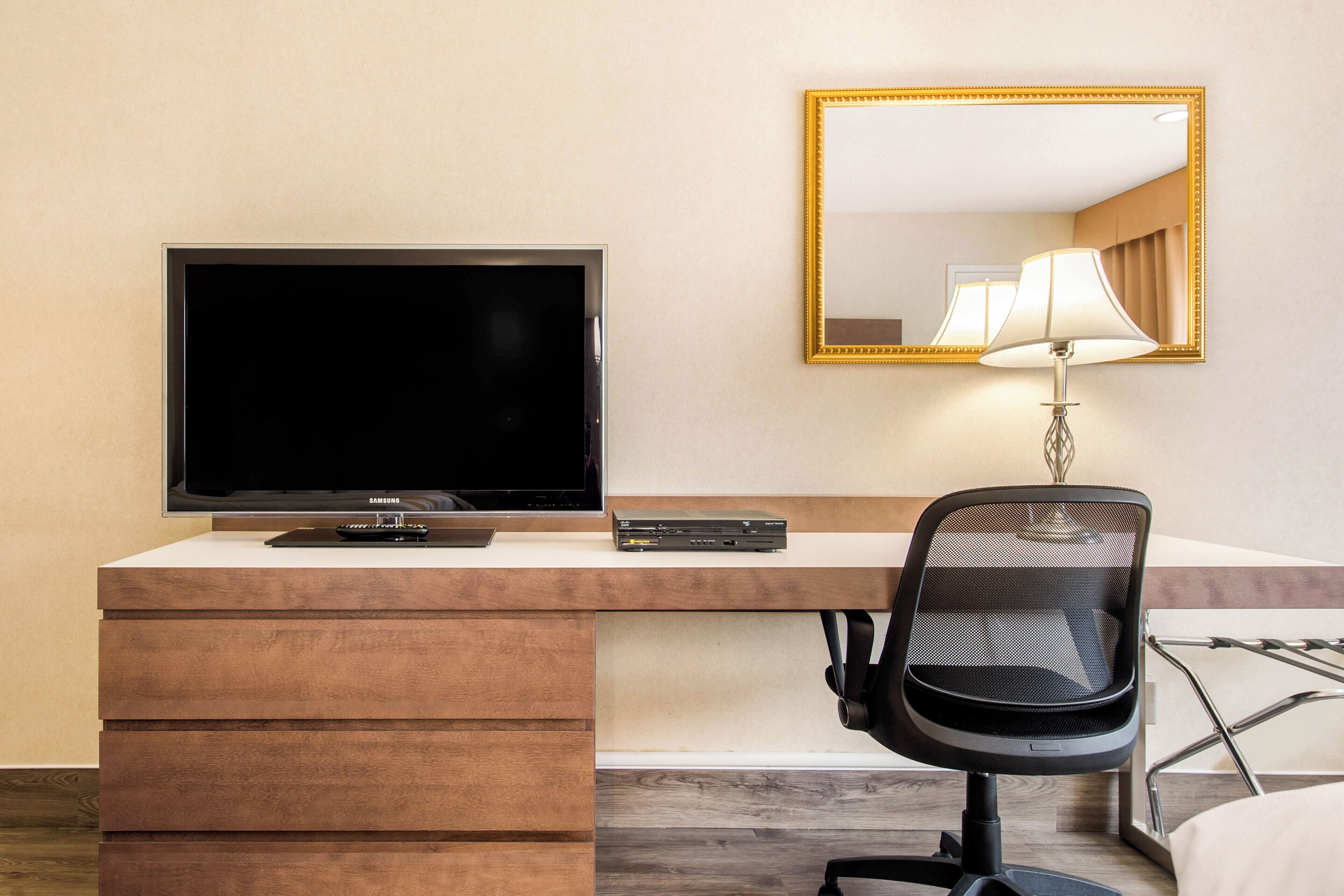 Habitación de hotel con televisión de pantalla plana, escritorio con lámpara y espejo en la pared.