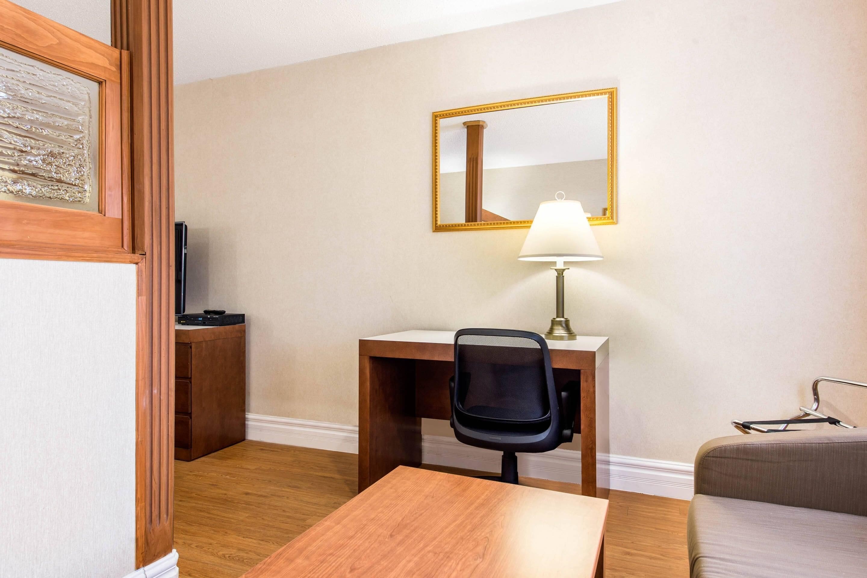 Habitación de hotel con un escritorio de madera, una silla, una lámpara, un espejo, un televisor y un sofá.