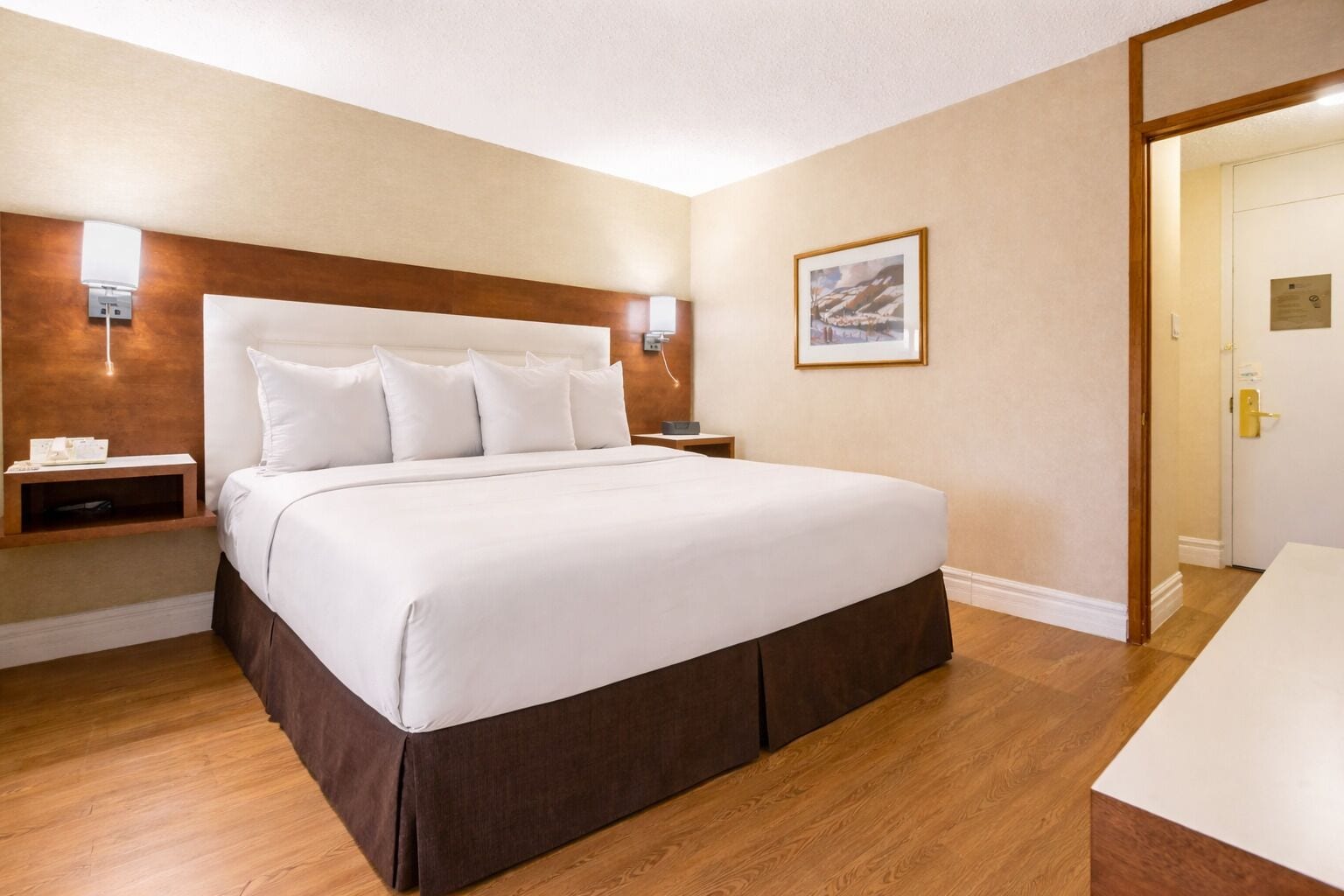 Una cama bien hecha en una habitación de hotel, con cabecera de madera, mesita de noche e imagen enmarcada en la pared.