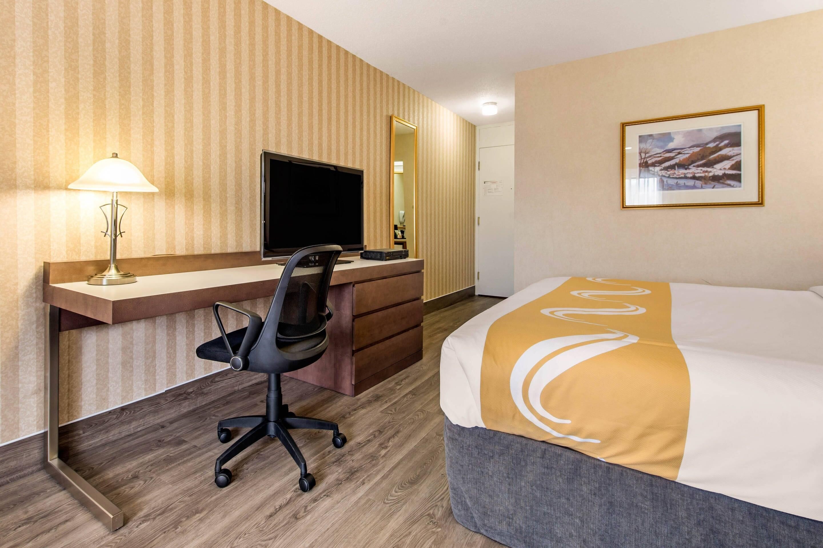 A hotel room with a wooden desk, a black office chair, a bed with a white and gold bedspread, a large mirror, and a framed picture on the wall.