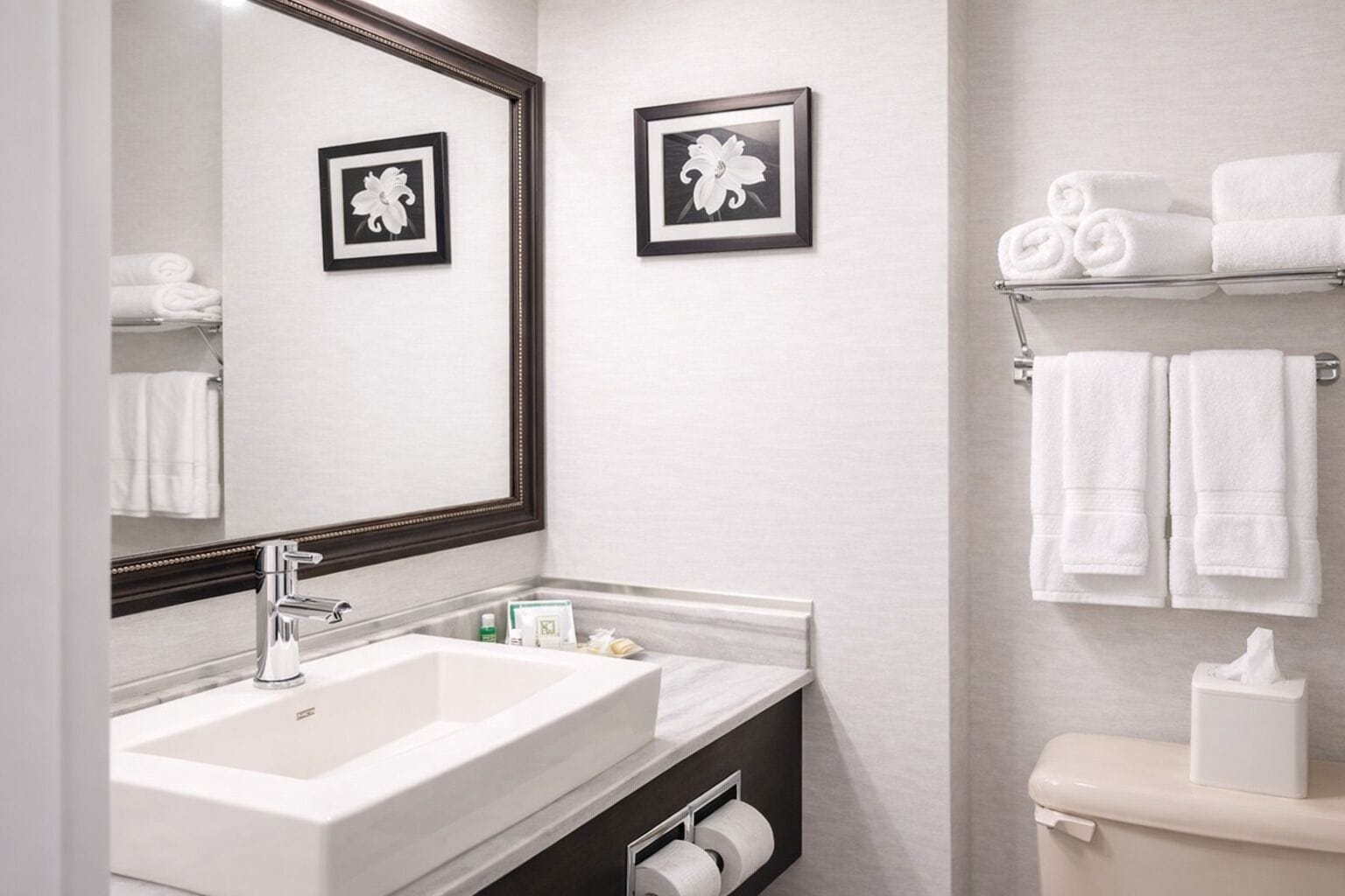 A modern bathroom with a large mirror, two framed floral pictures, a white sink, and a toilet with a towel rack above it.