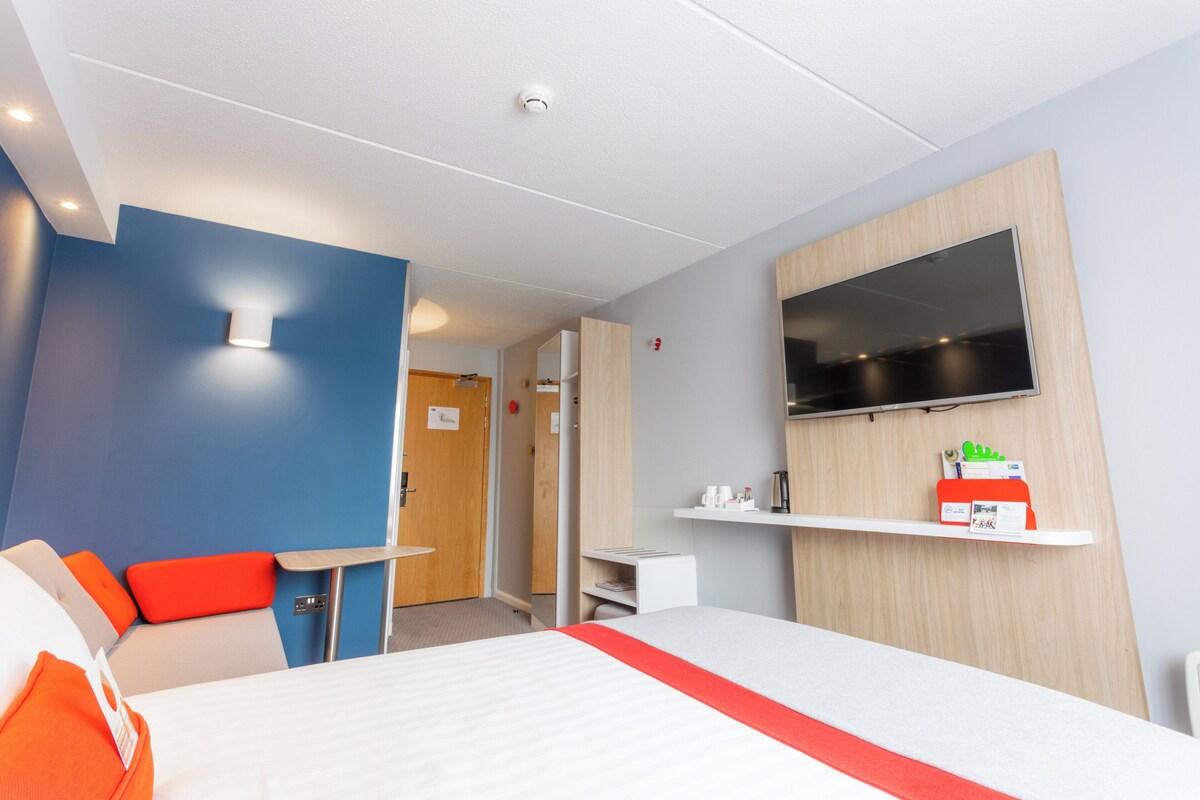 A hotel room with a bed, a desk, a chair, a TV, and a shelf with amenities.