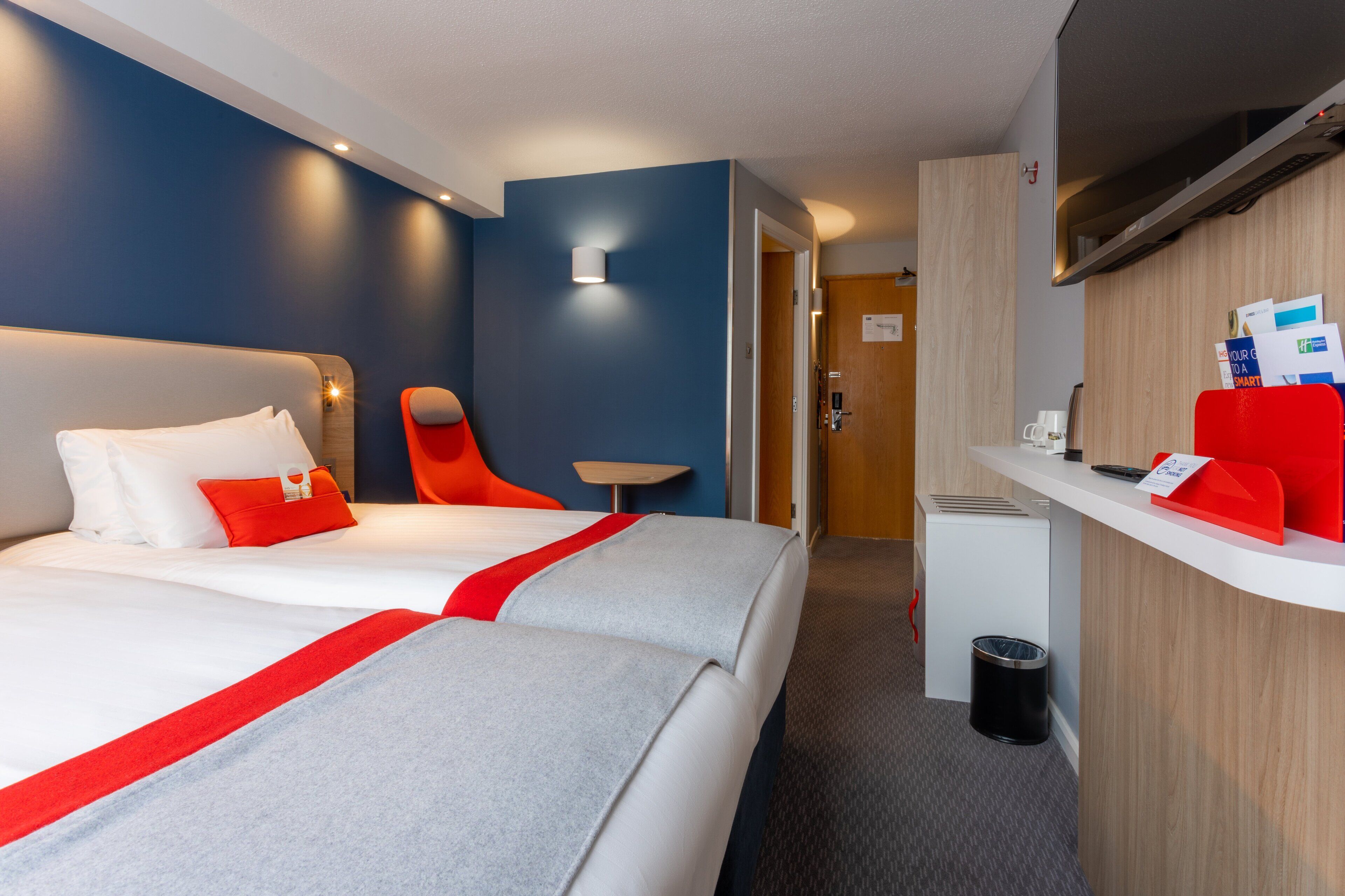 A hotel room with two beds, a desk, a chair, a small table, and a trash bin.