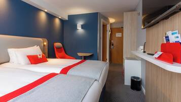 A hotel room with two beds, a desk, a chair, a small table, and a trash bin.