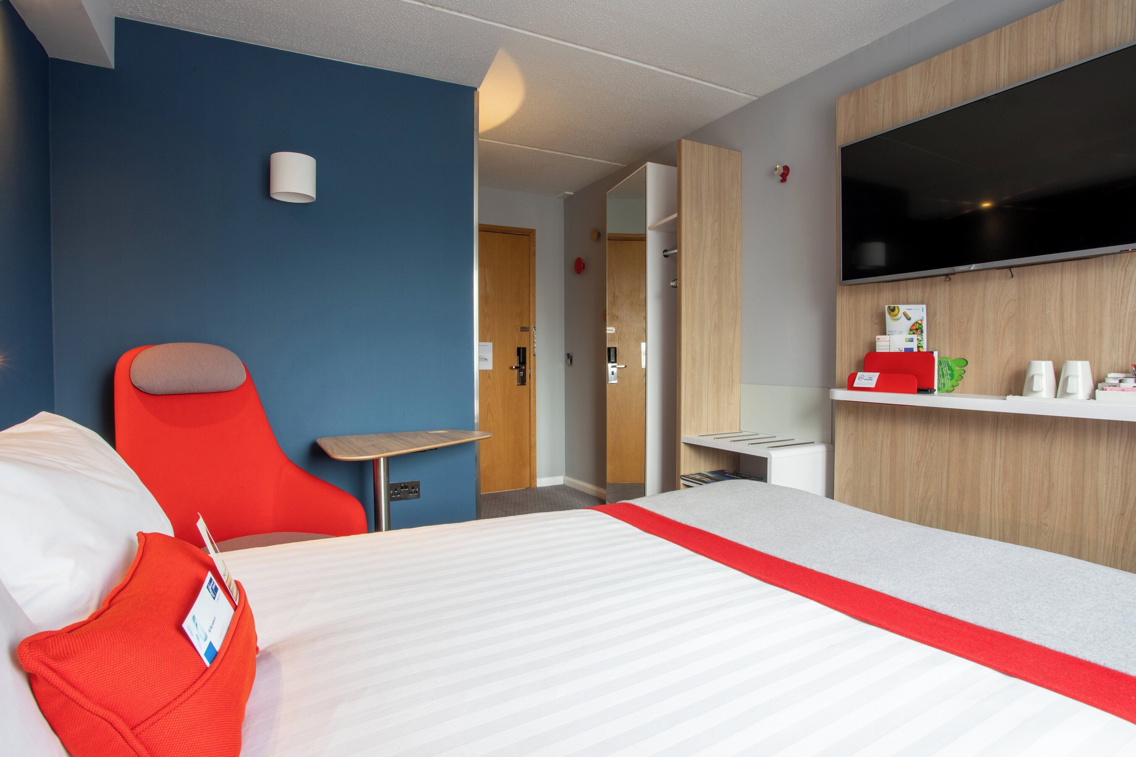 A hotel room with a bed, a red chair, a small table, a TV, and a wooden cabinet with a shelf.