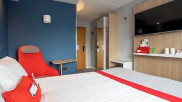A hotel room with a bed, a red chair, a small table, a TV, and a wooden cabinet with a shelf.