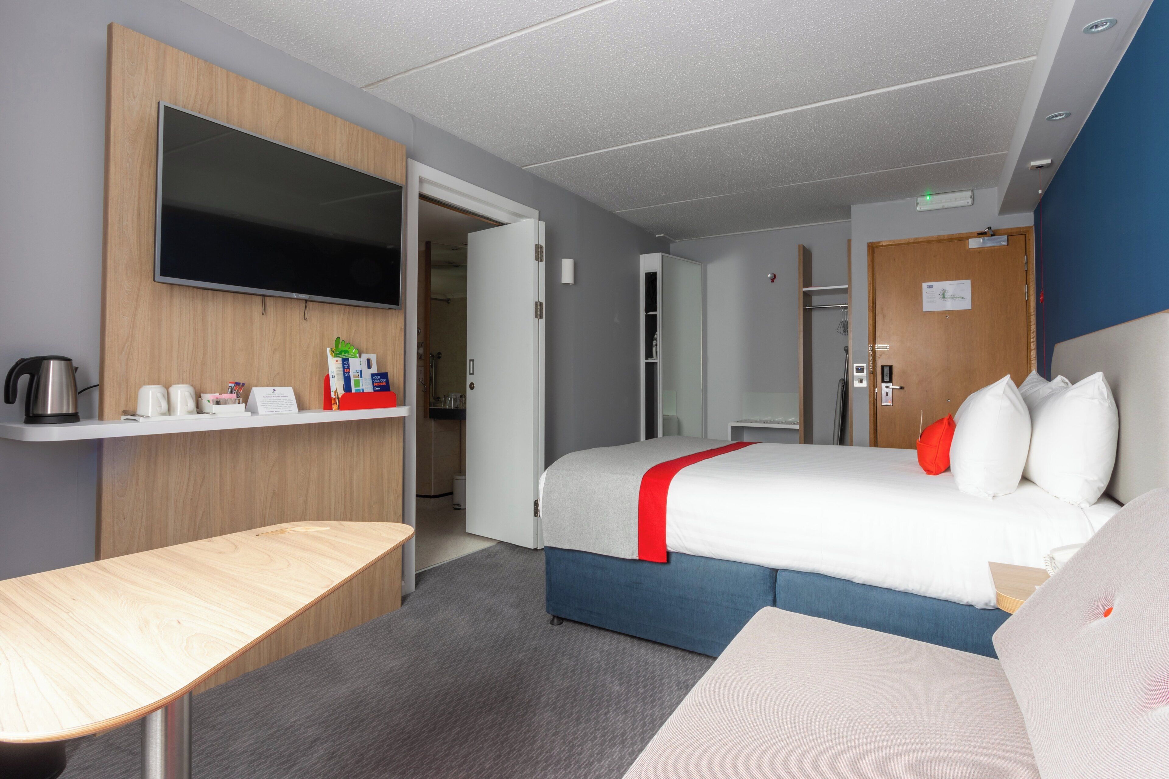 Une chambre d’hôtel avec un lit, un téléviseur fixé au mur, un bureau et une salle de bain visible à travers une porte ouverte.