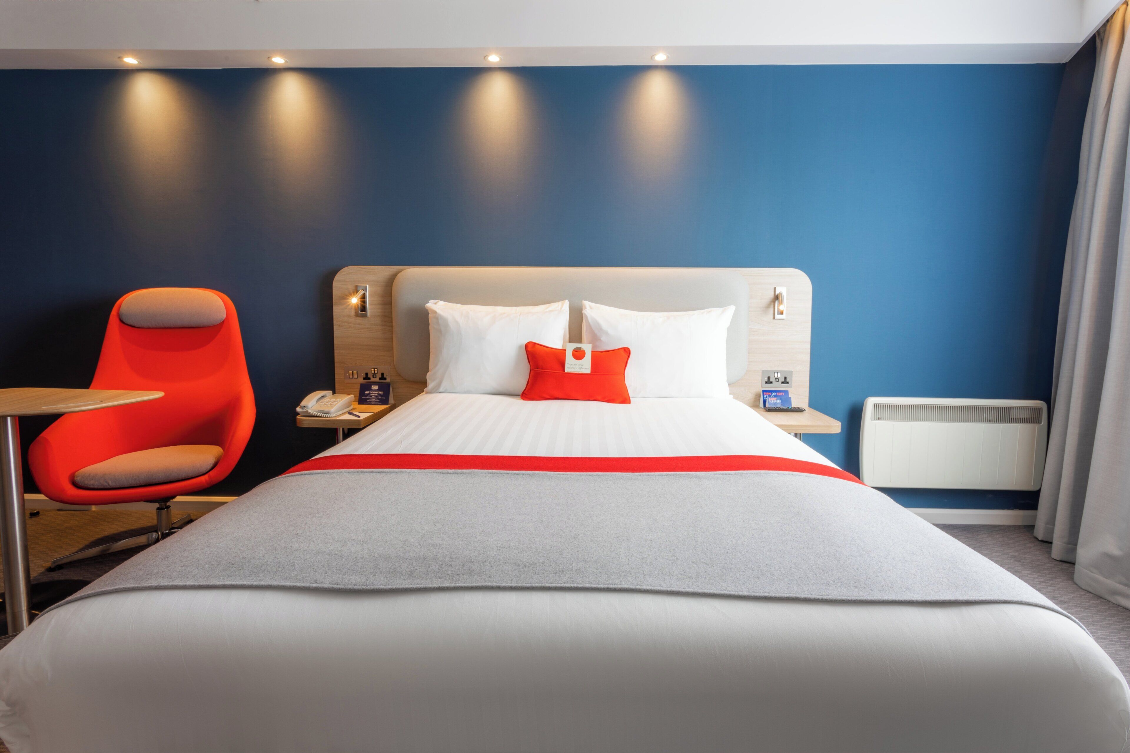A hotel room with a large bed, a red chair, a bedside table with a phone, and a nightstand with a lamp.
