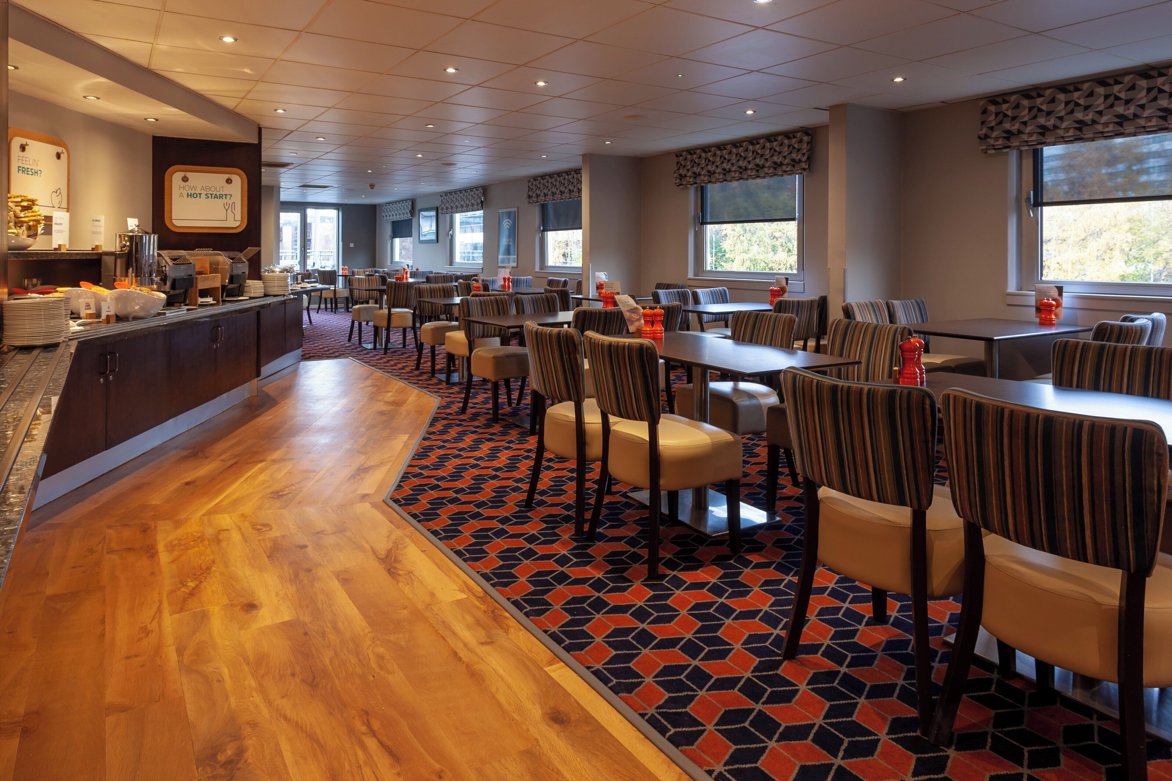 A hotel dining area with multiple tables and chairs, a buffet counter, and large windows providing natural light.
