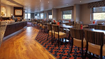 A hotel dining area with multiple tables and chairs, a buffet counter, and large windows providing natural light.