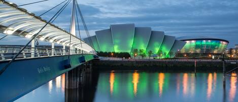 A waterfront view of a modern architectural structure with a bridge, calm water, and a stadium.