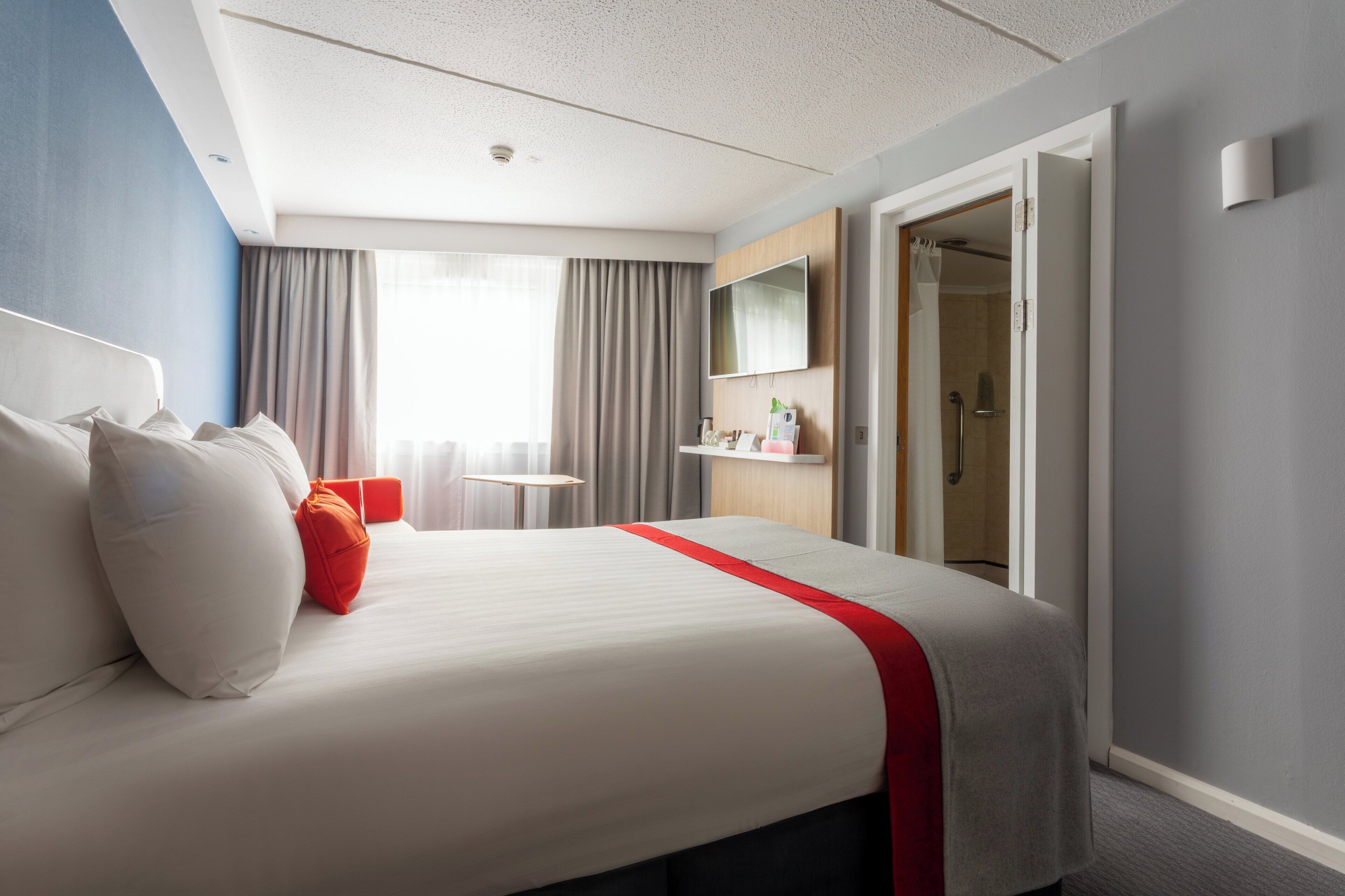 Ein Hotelzimmer mit einem großen Bett, einem kleinen Tisch, einem Fernseher und einem Duschbereich.