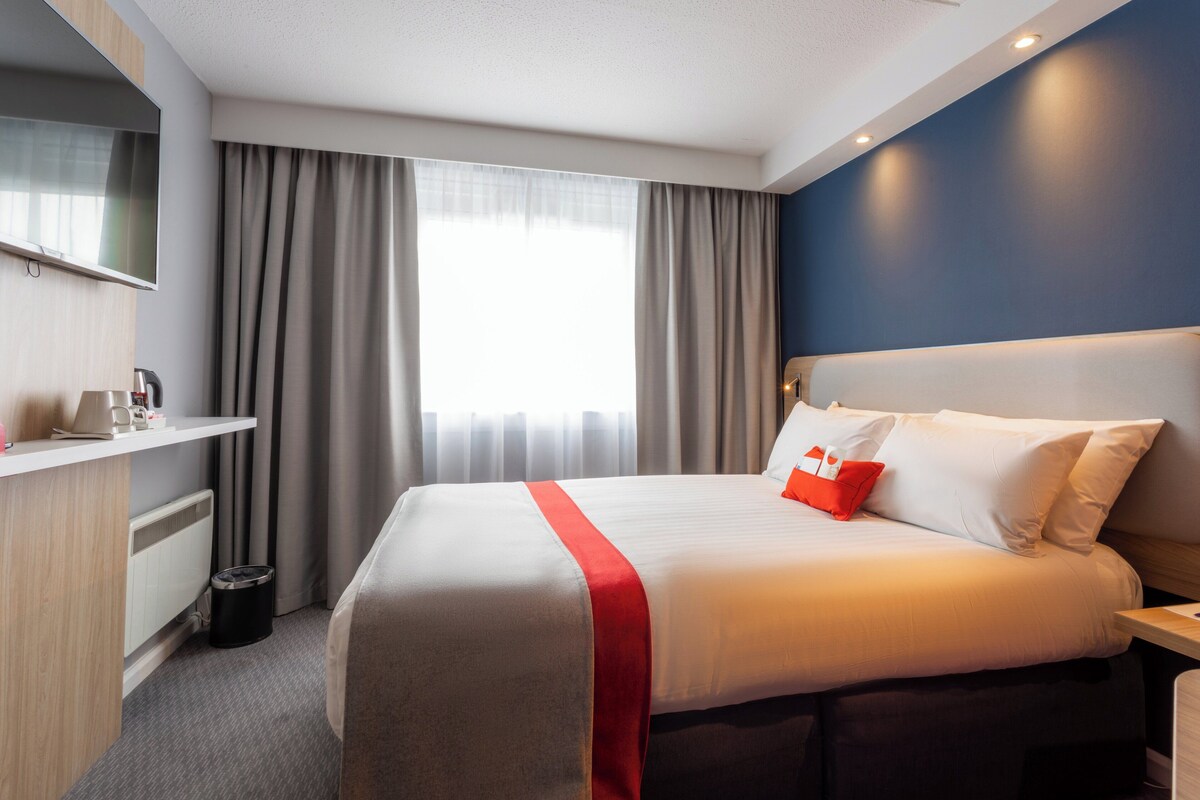 A hotel room with a bed, a bedside table, a TV mounted on the wall, and a window with curtains.