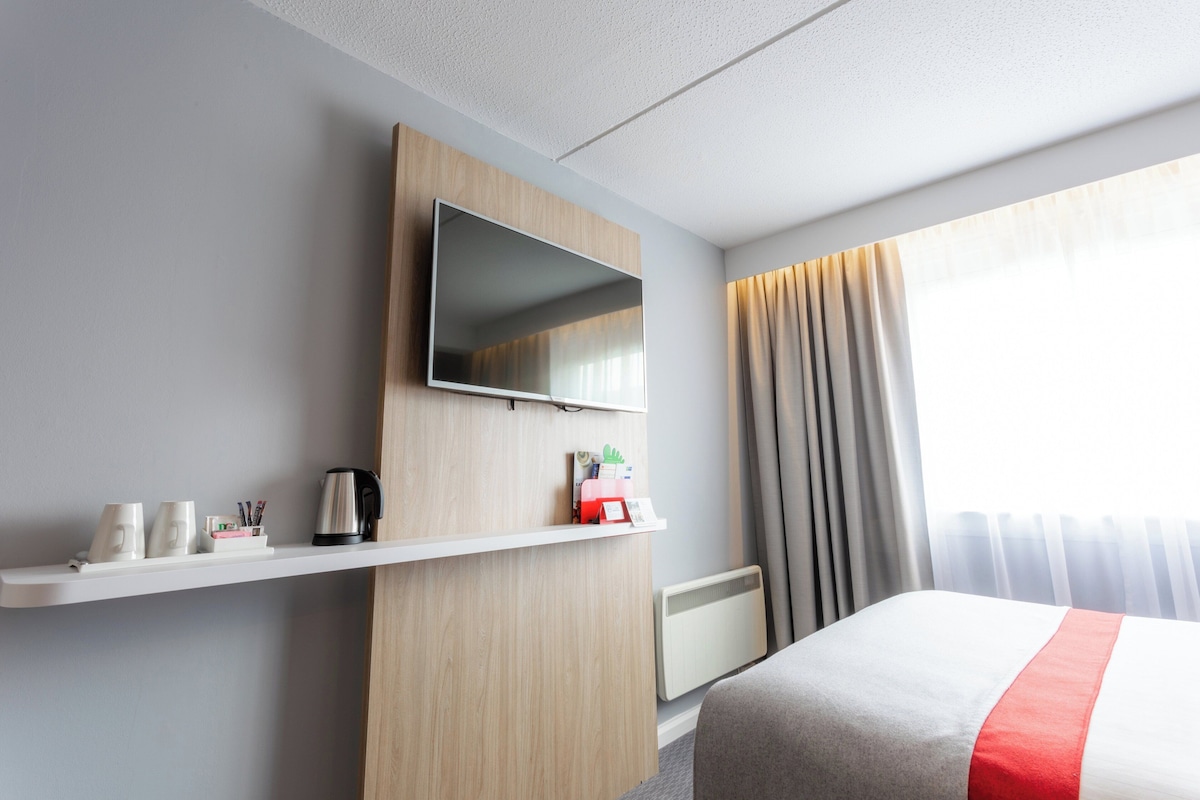 Habitación de hotel con cama, televisor montado, repisa con artículos de aseo, radiador y ventana con cortinas.