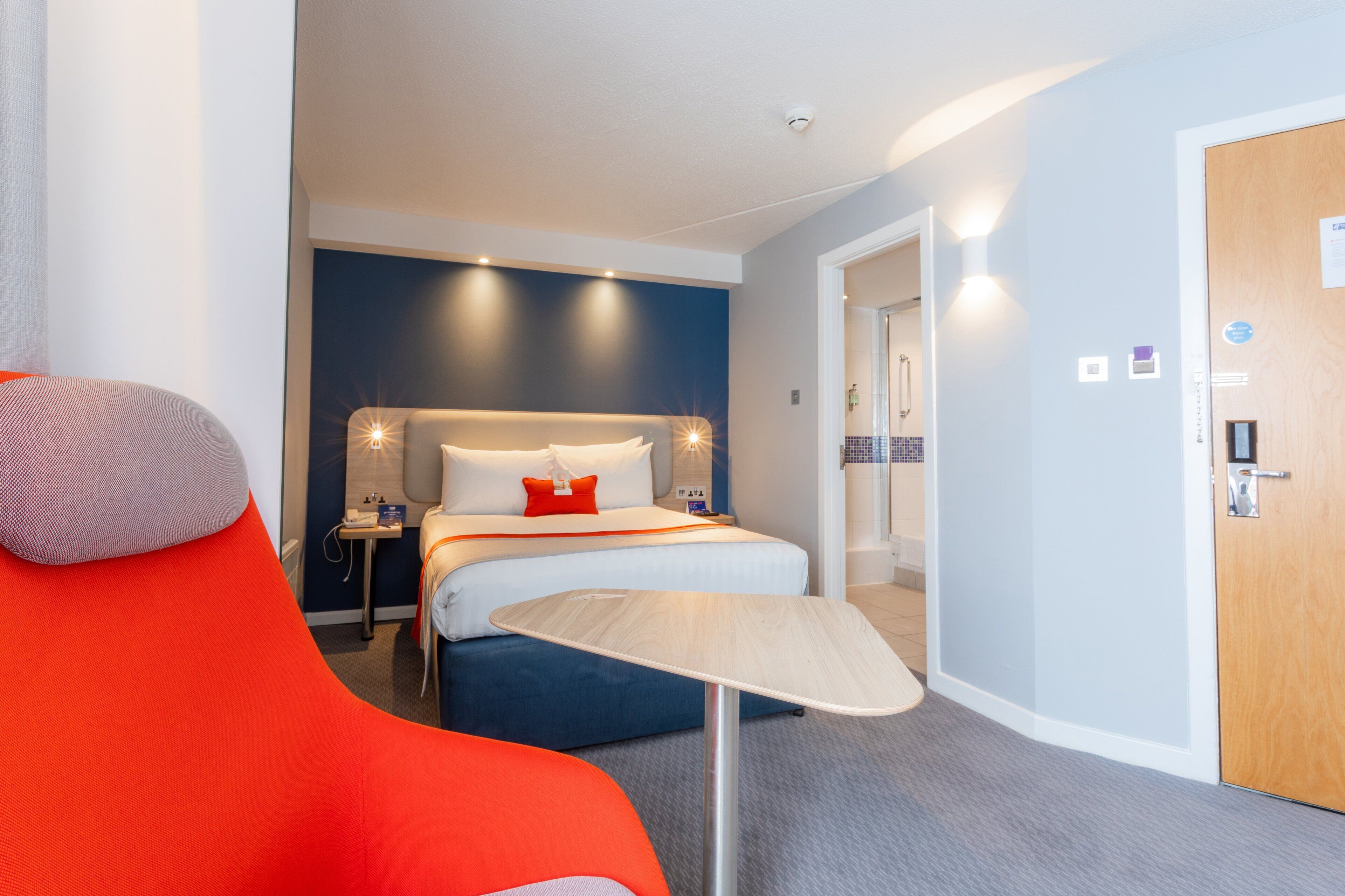 A modern hotel room with a bed, bedside table, and a small table with a chair.