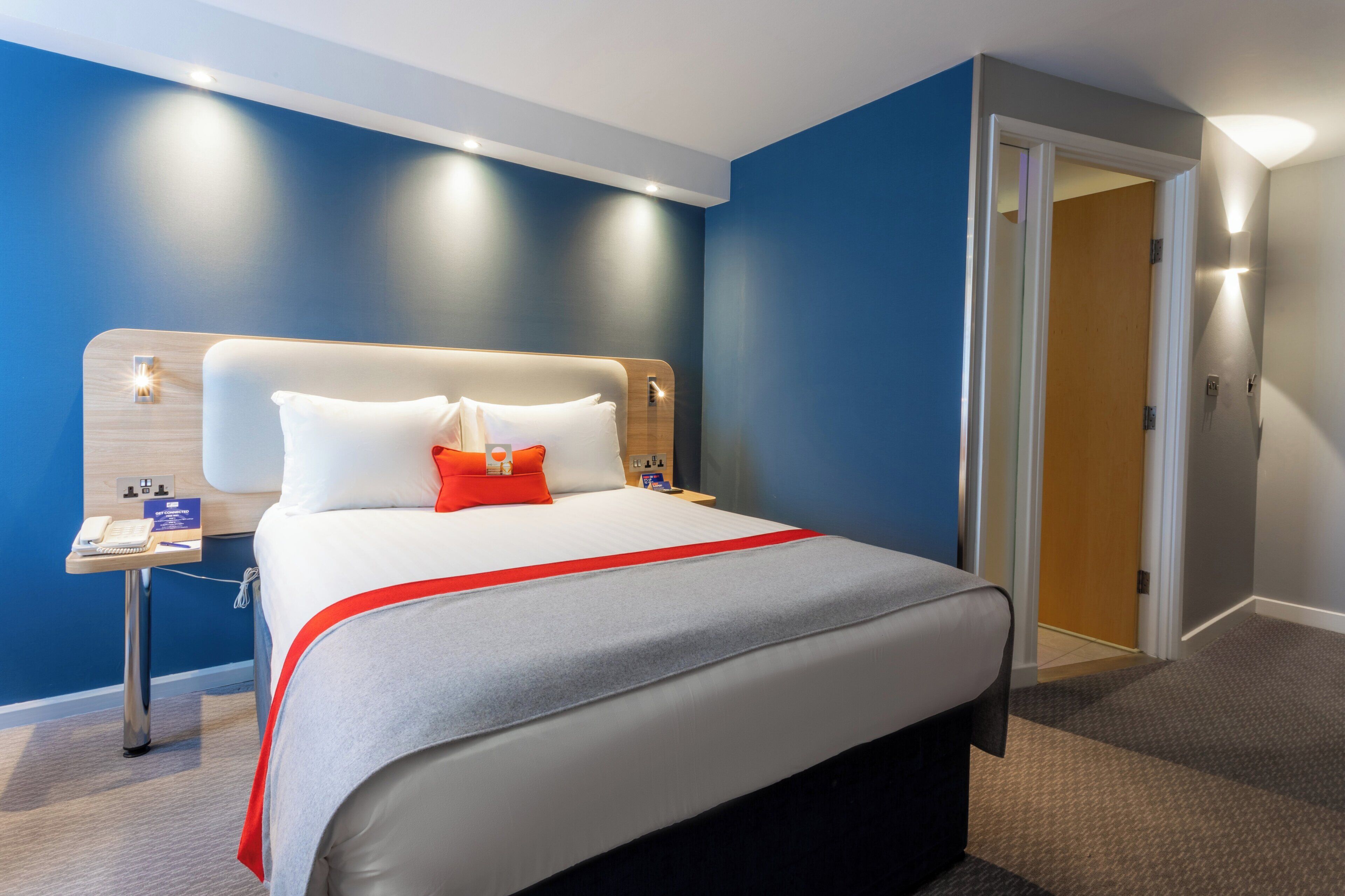 Een hotelkamer met een bed, nachtkastje en een deur die leidt naar een andere kamer.