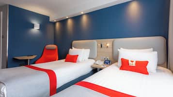 Une chambre d’hôtel avec deux lits, chacun agrémenté d’un coussin rouge, une table de chevet avec un téléphone et un panneau, et une seule chaise.
