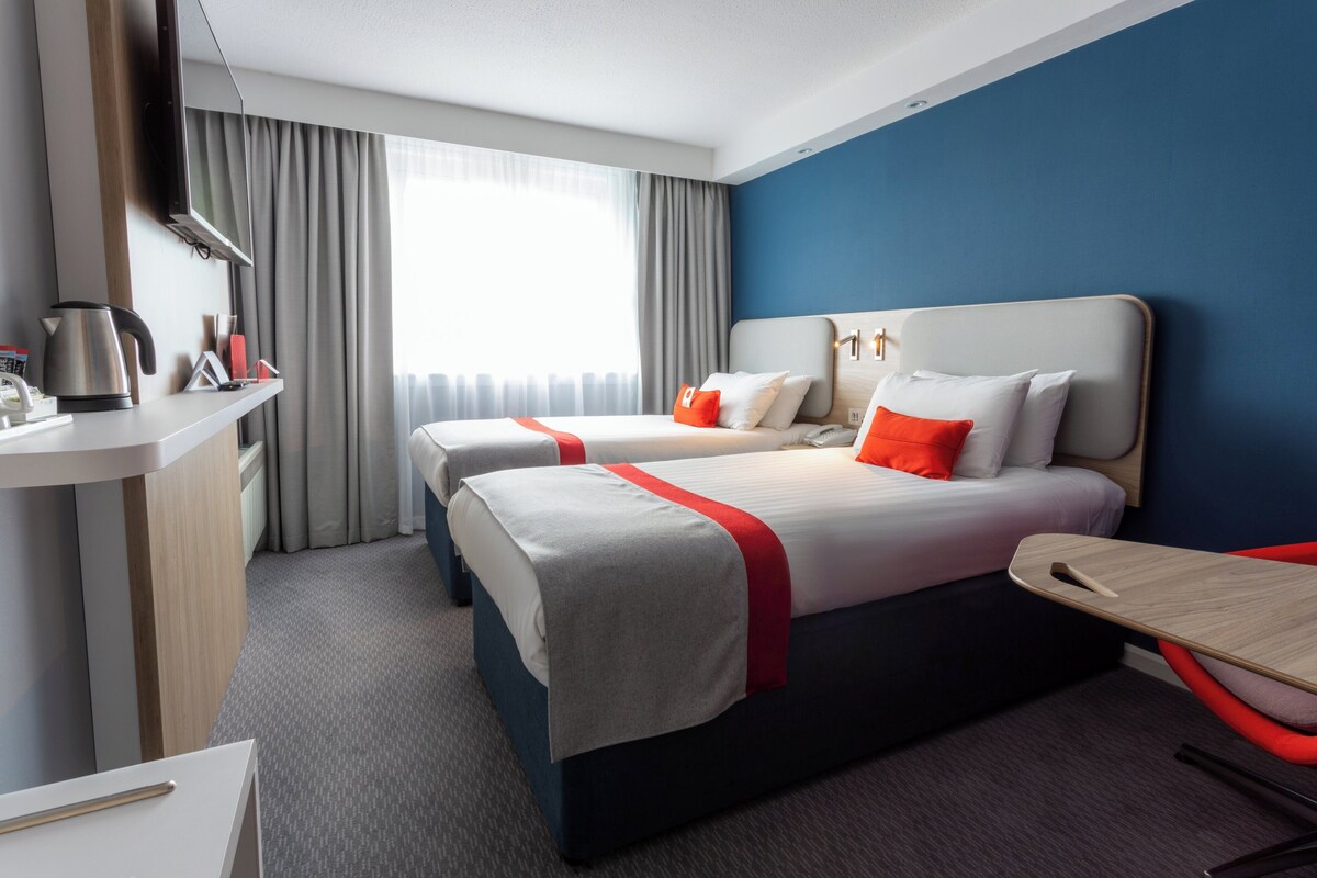 A hotel room with two beds, a desk, a chair, a coffee maker, and a window with curtains.