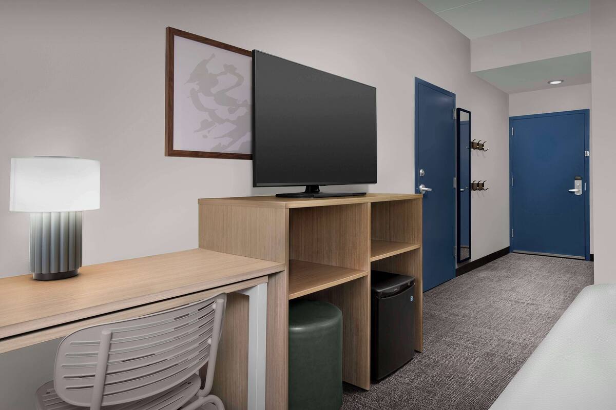 A hotel room with a desk, chair, TV, lamp, trash bin, and a bed.