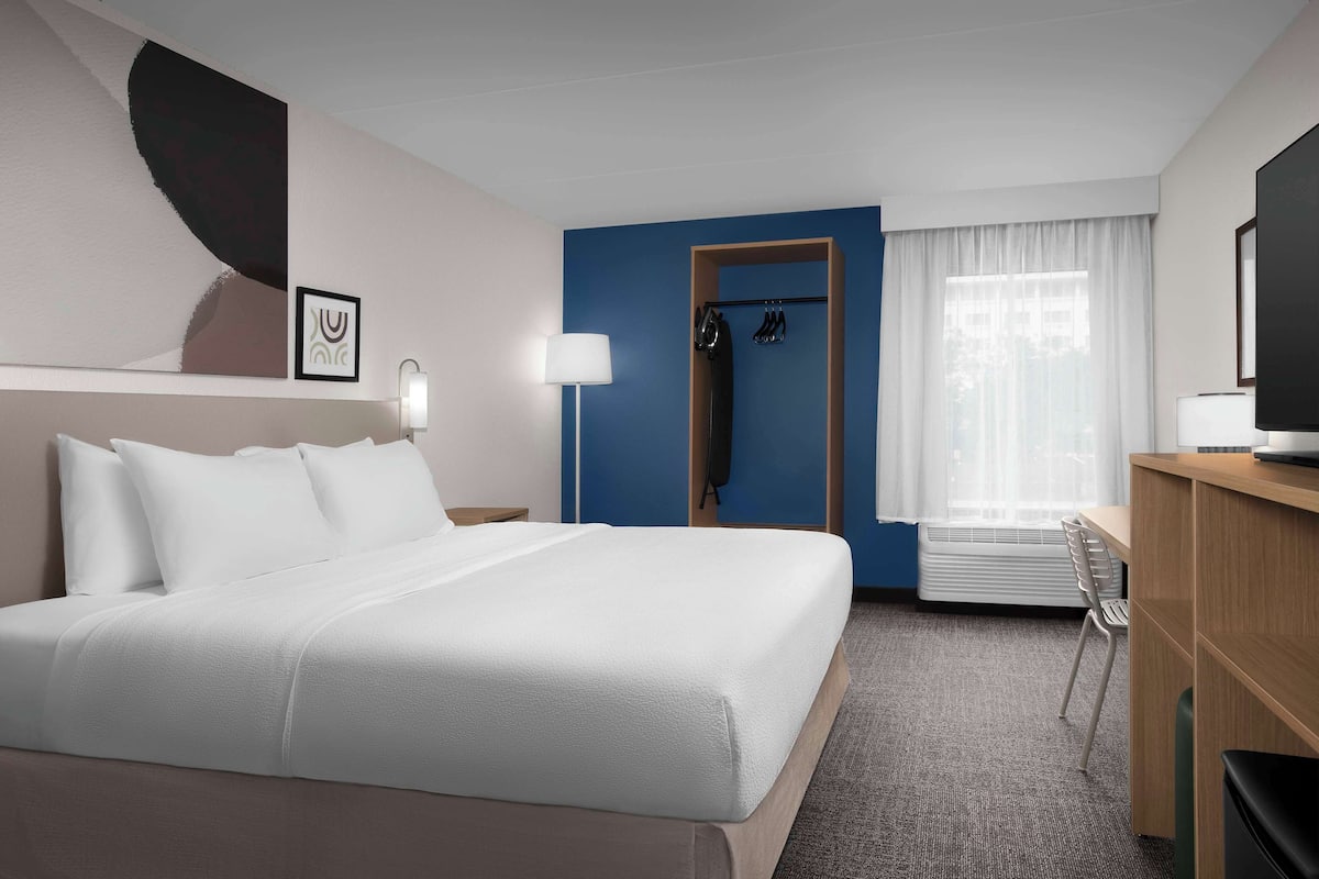 A hotel room with a large bed, a desk, a chair, a coat rack, and a television.