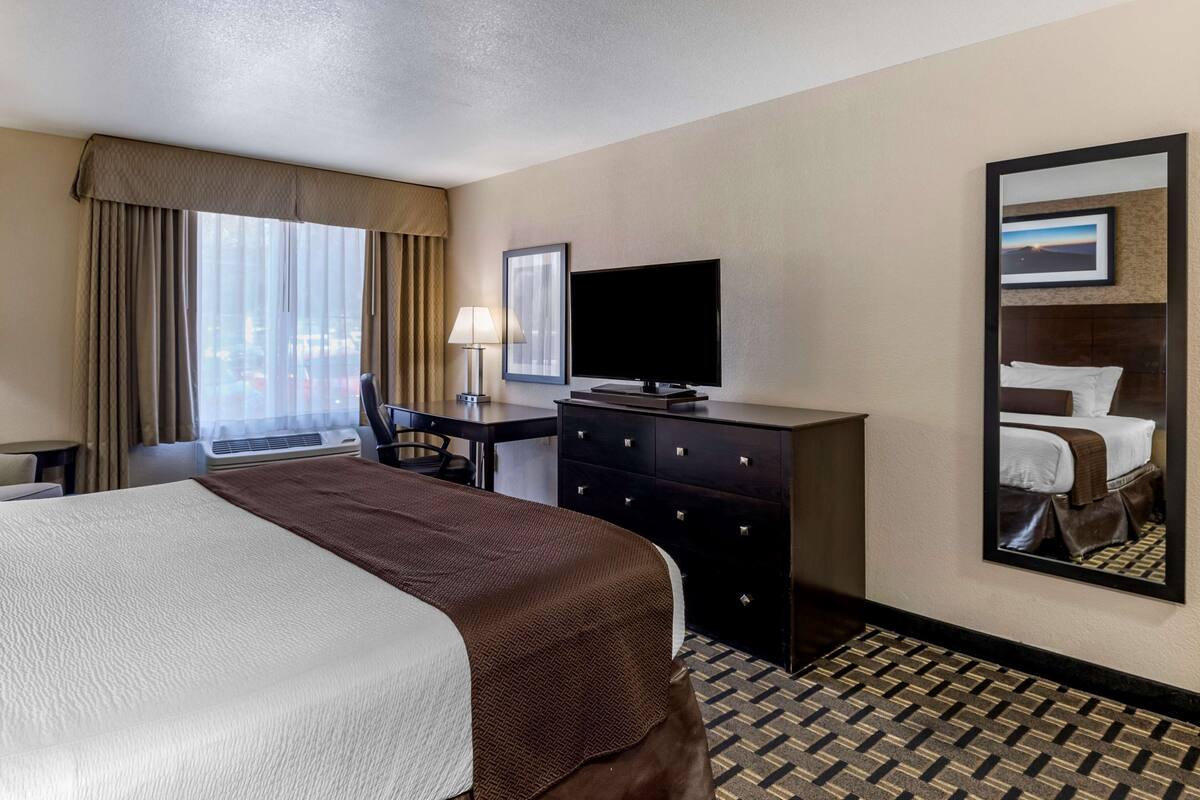 Ein Hotelzimmer mit einem großen Bett, einem Schreibtisch mit Stuhl, einem Flachbildfernseher, einem Schminktisch und einem großen Spiegel, der das Bett reflektiert.