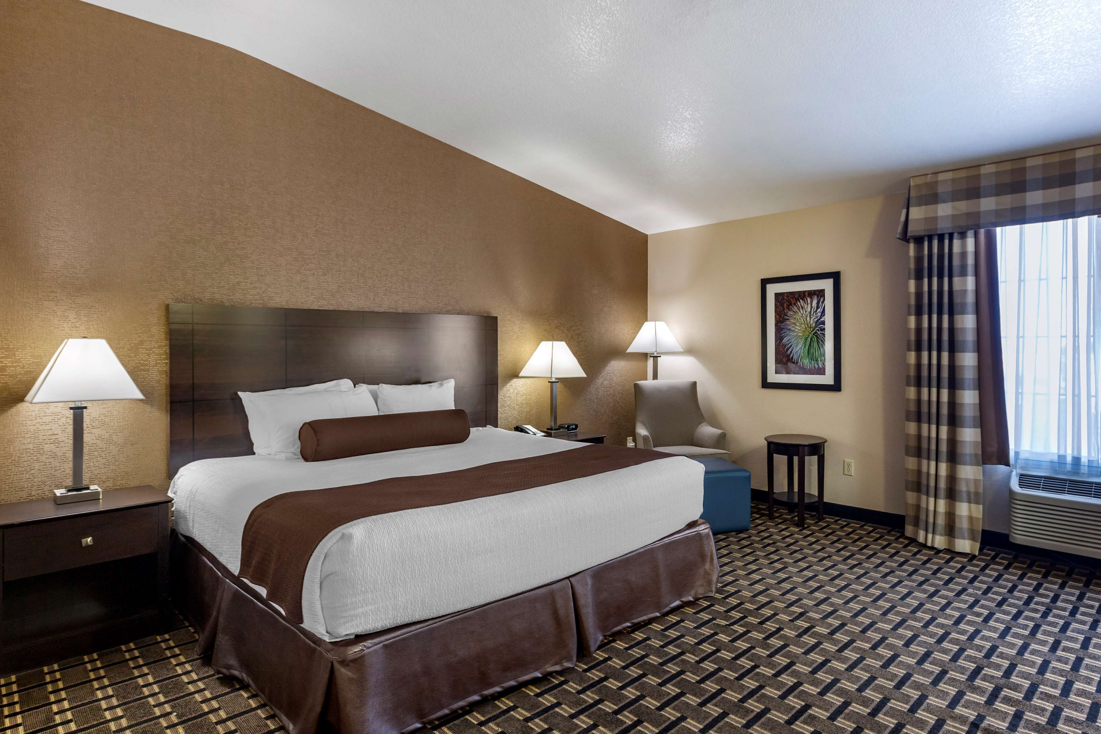 Una habitación de hotel con una cama grande, dos lámparas de noche, una silla, una mesita y una ventana con cortinas.