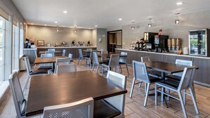 Un espace repas moderne avec plusieurs tables et chaises, un coin cuisine équipé d’appareils électroménagers et d’un réfrigérateur, ainsi qu’une grande fenêtre laissant entrer la lumière naturelle.