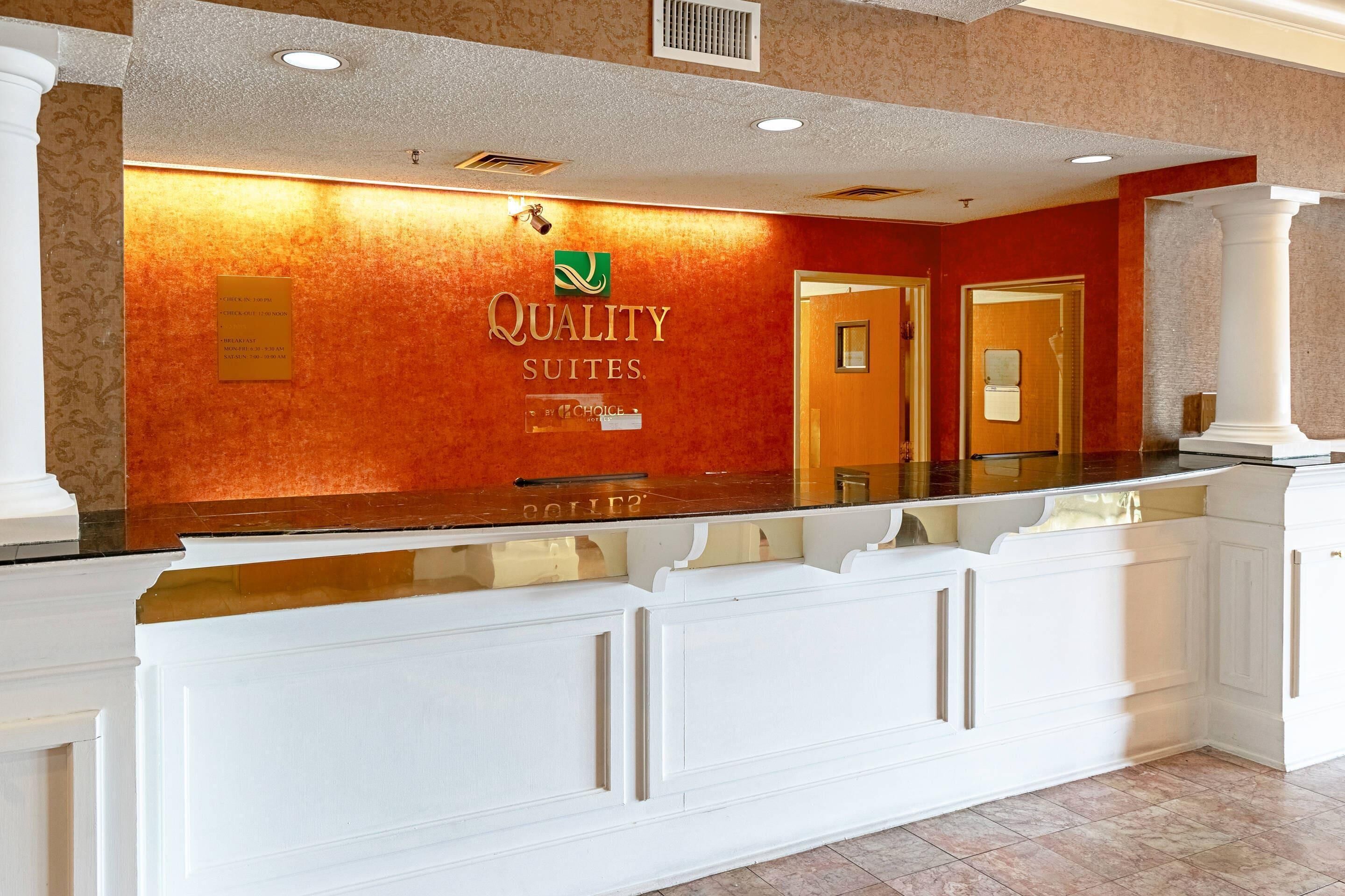 Una reception con una insegna che recita 'Quality Suites' e un logo sopra. Si notano due porte sullo sfondo.